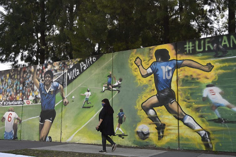 Una mujer vestida de negro pasa frente a una pared con imágenes de Diego Maradona corriendo.