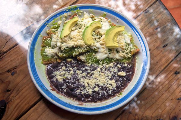 Chilaquiles con salsa verde, queso cotija, crema, cebolla y aguacate, acompañados de frijoles negros refritos