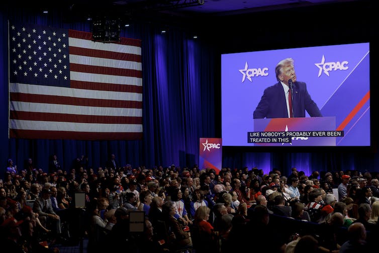 Una gran multitud de personas mira hacia una pantalla que muestra a un hombre blanco con un traje oscuro. Junto a la pantalla hay una gran bandera estadounidense.