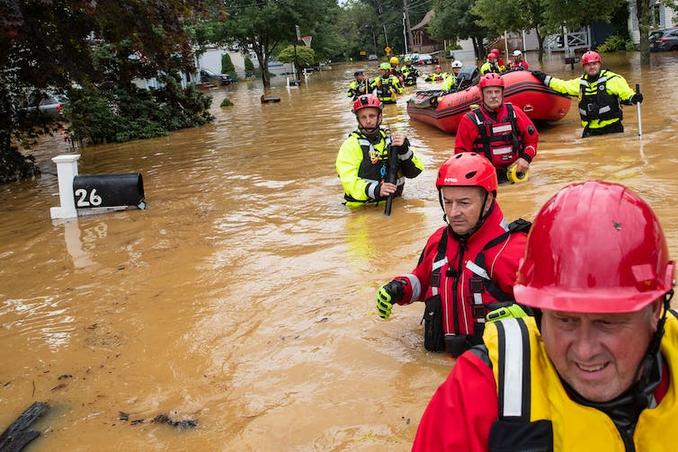 Una fila de trabajadores de rescate con chalecos brillantes y cascos camina con agua hasta la cintura en una calle inundada, tirando de una balsa. El agua llega hasta el buzón por el que pasan.