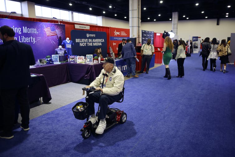 Un hombre en silla de ruedas pasa frente a una cabina en una sala de convenciones que dice 
